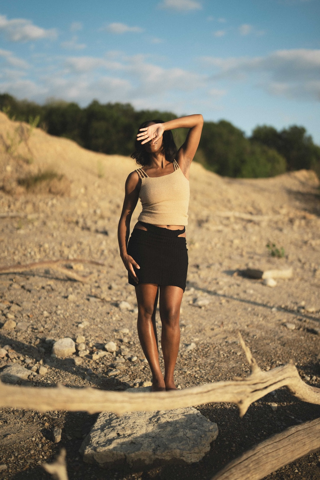 Woman standing on a rocky landscape with trees in the background wearing a knit beige tank top and black skirt 