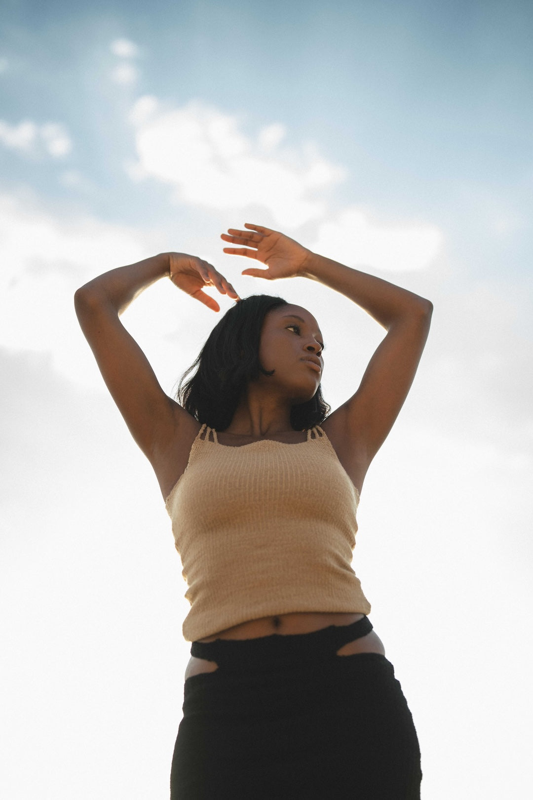 Woman with arms raised against a bright sky in camel knit tank top 