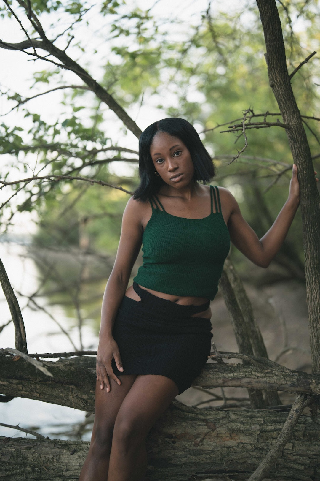 Woman in a green top and black skirt sitting on a tree branch by a body of water.