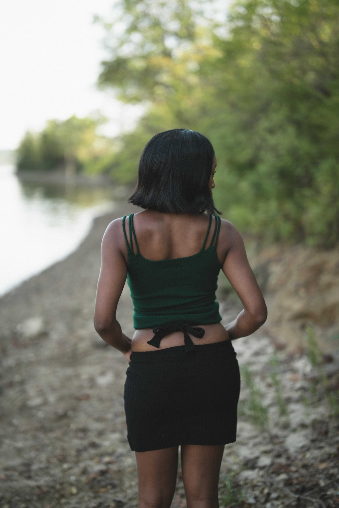 Person wearing a green tank top and black skirt standing by a body of water with trees in the background.