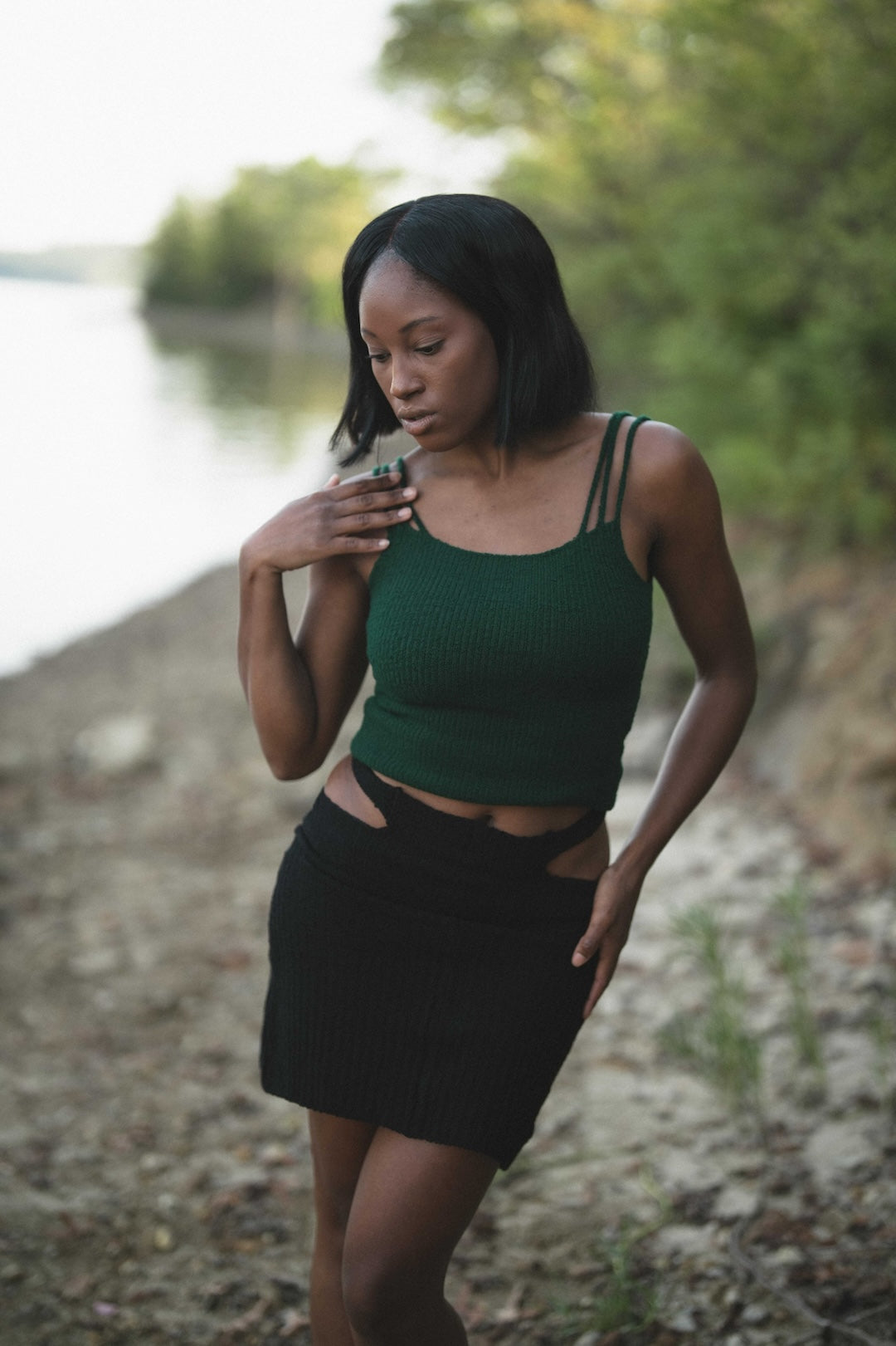 Woman in green top and black skirt standing by a body of water with trees in the background