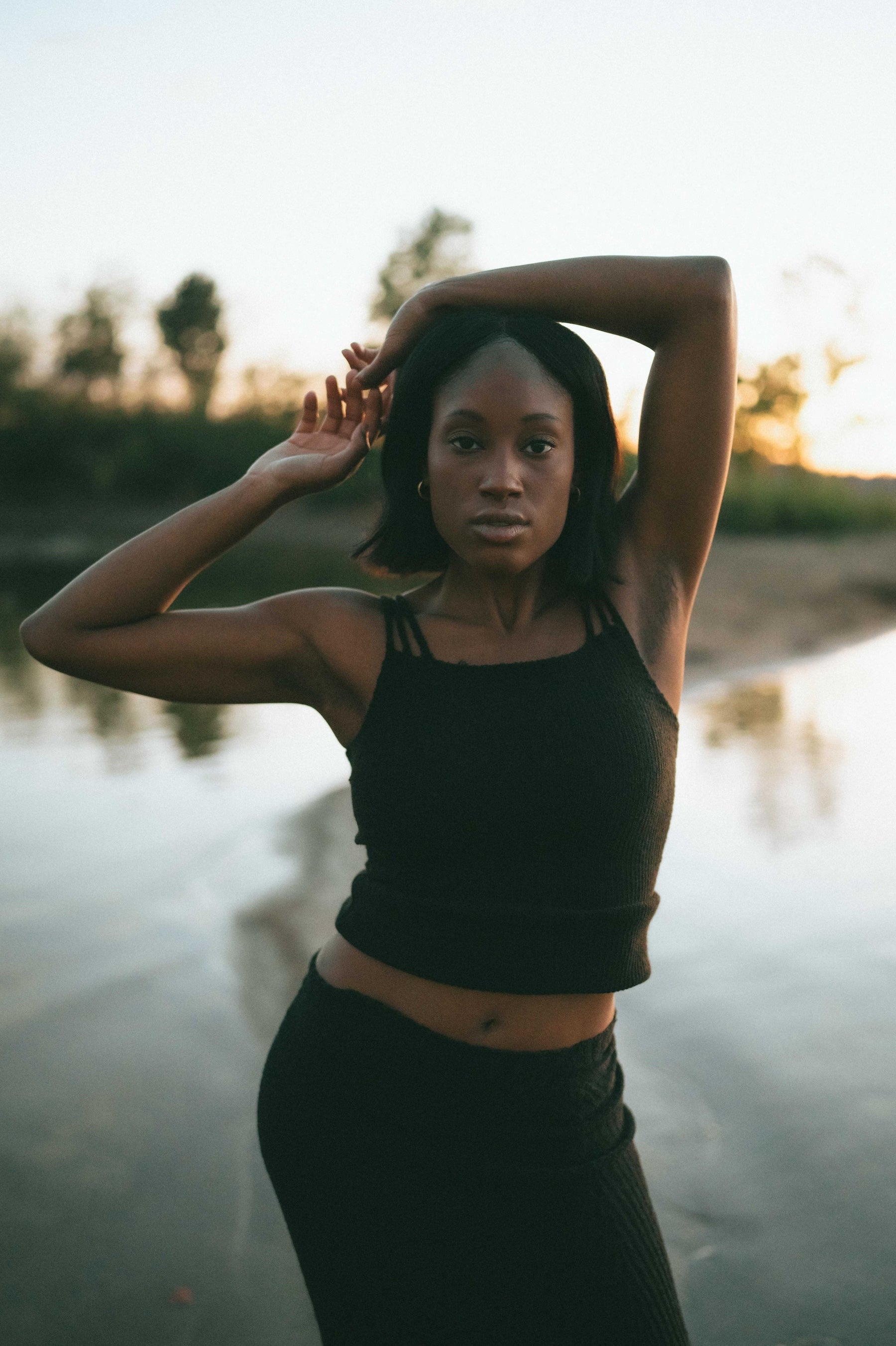 Woman in black tank top and skirt standing by a body of water with sunset in the background