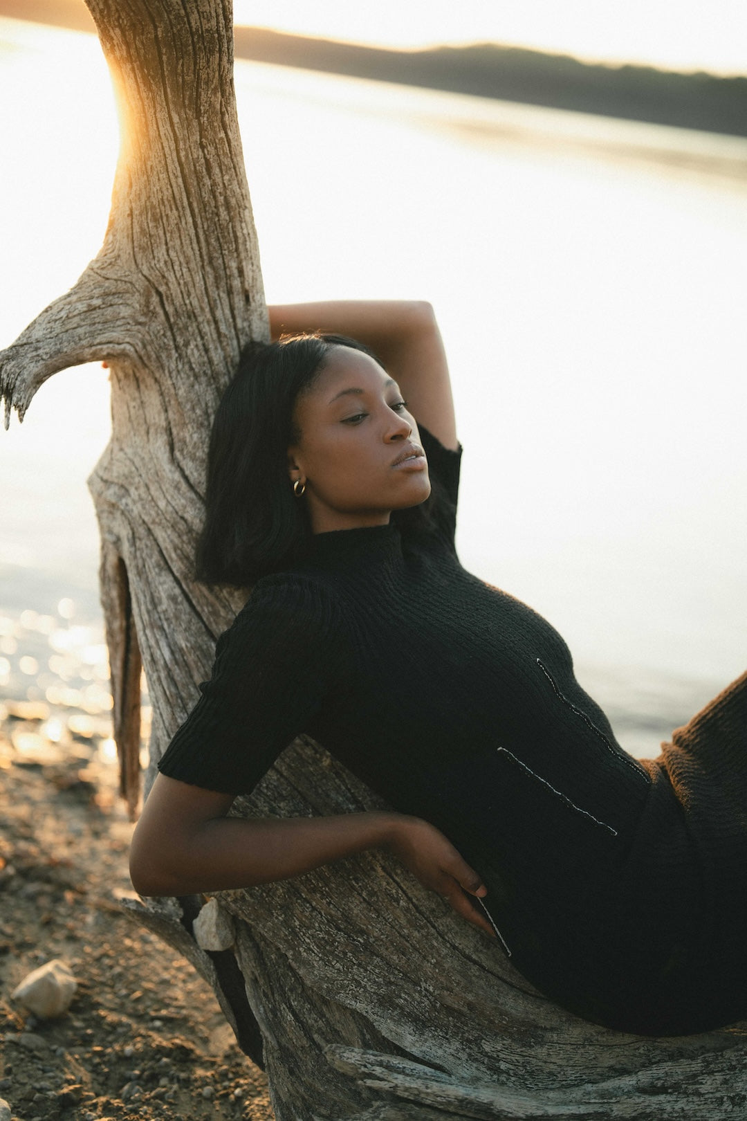 Woman in a black knit dress leaning against a tree by a body of water at sunset.