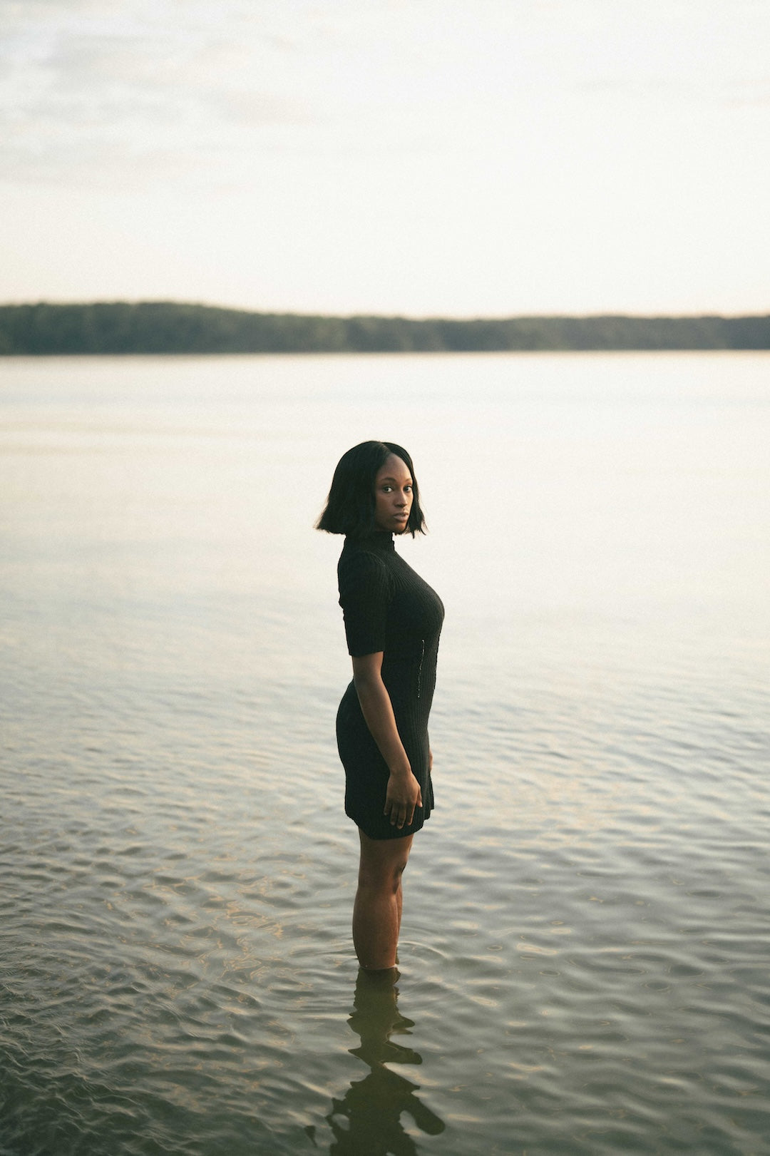 Woman standing in a body of water wearing a knit dress with a sunset in the background